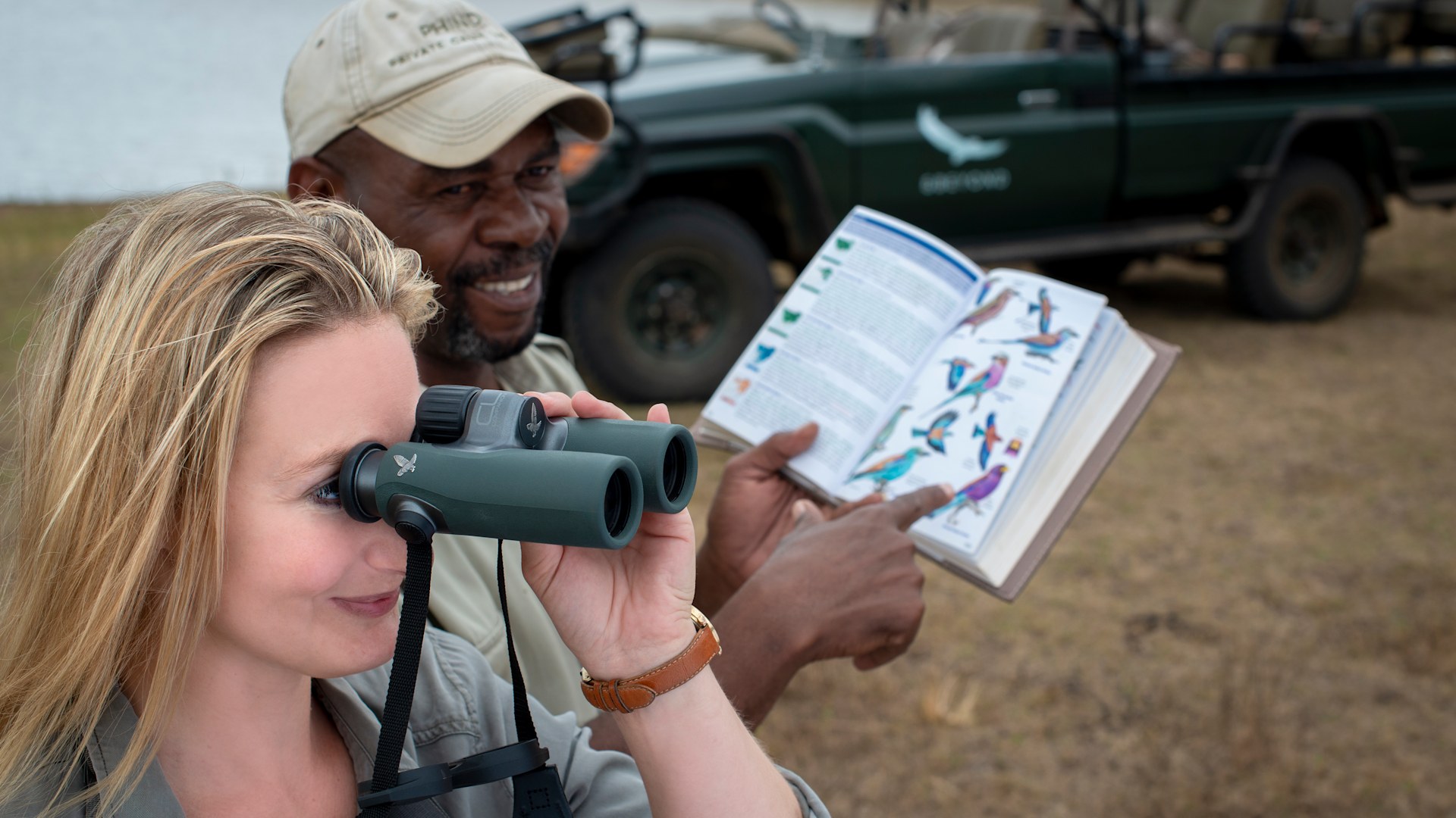 Safari birdwatchers using binoculars and a bird field guide on a game drive in Africa