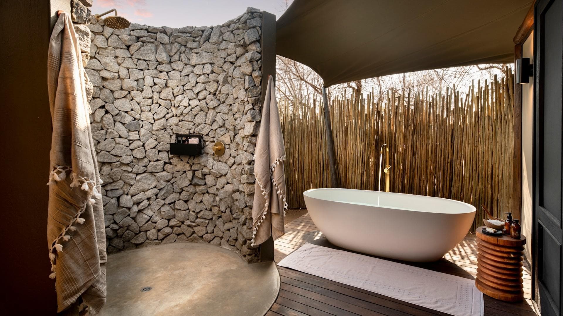 A luxury outdoor bath at a tented safari camp — stone walls, brass fittings, and the African bush beyond the bamboo screen