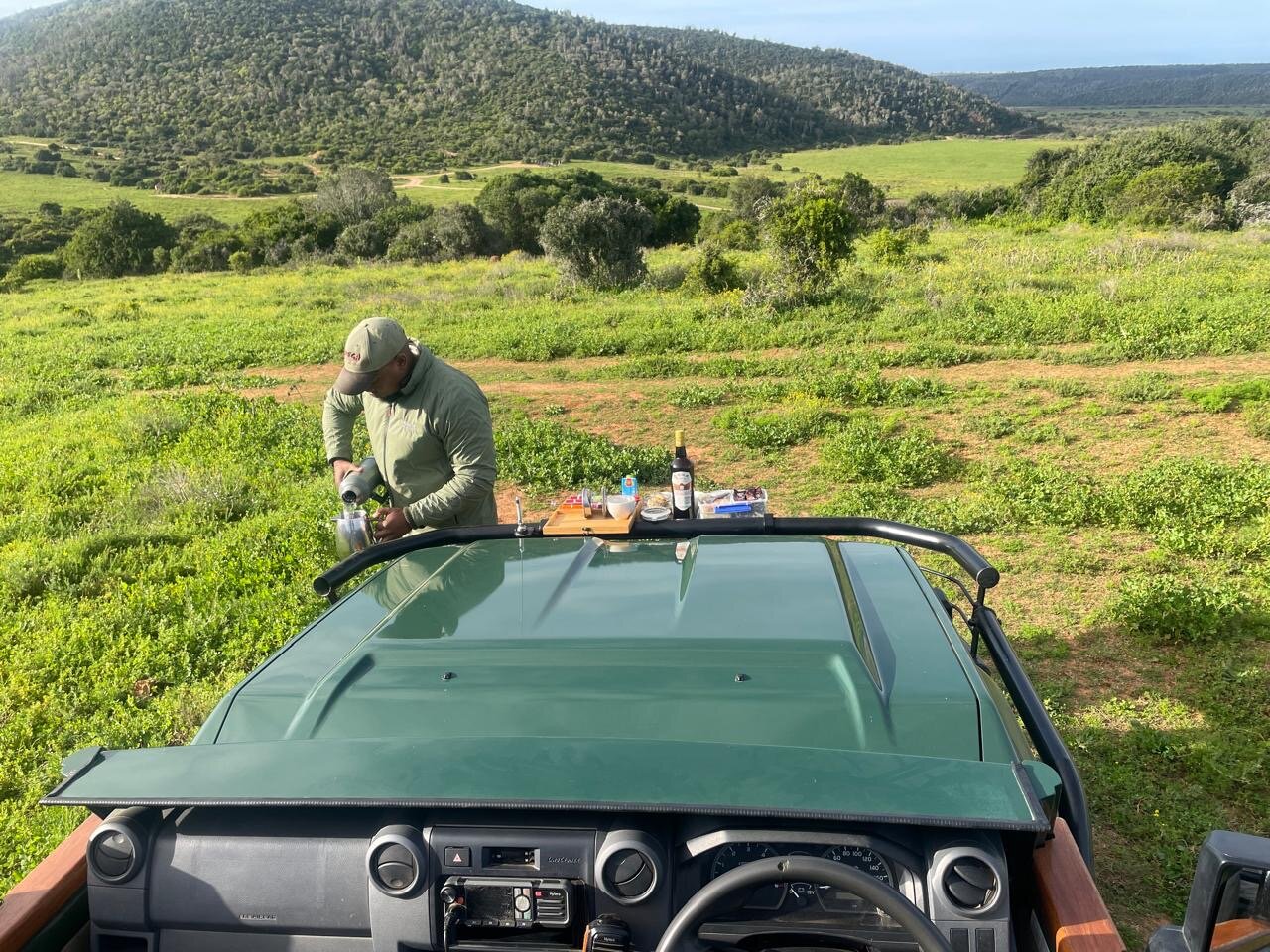 Our ranger pouring sundowner drinks laid out across the vehicle bonnet, the green Kariega valley stretching out behind