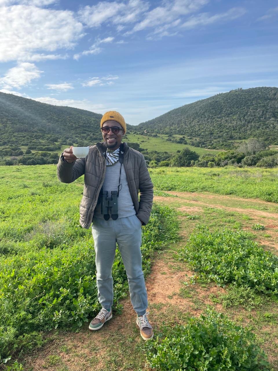 One of the founders smiling and holding out a cup of freshly brewed bush coffee, green Kariega hills rolling out behind