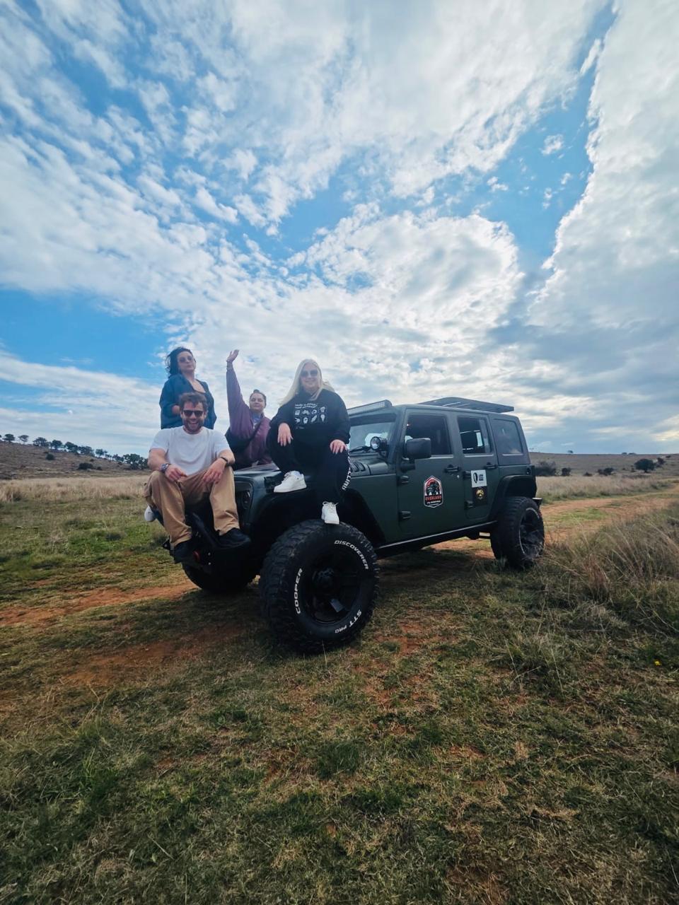 A group of friends sitting on the hood of a safari jeep at the Lion and Safari Park, big smiles and arms up