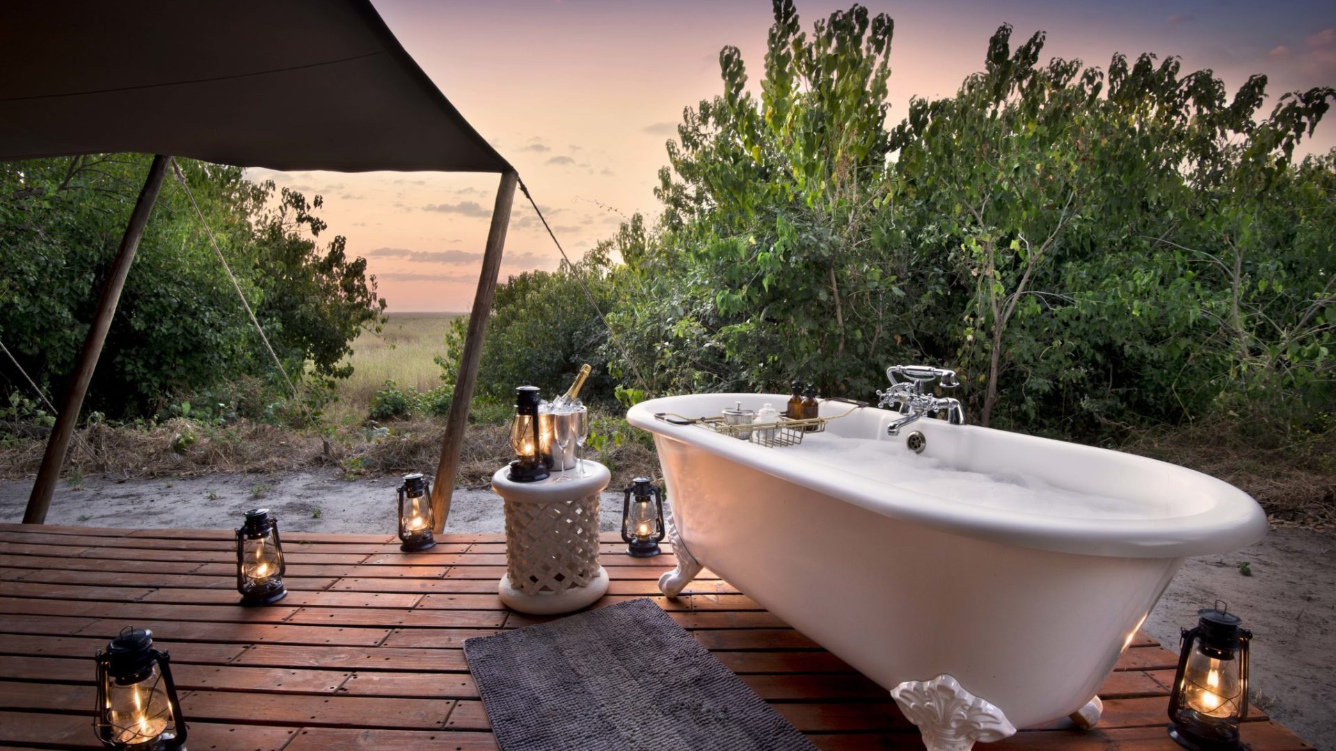 A luxury outdoor bath on a private wooden deck overlooking the African bush at sunset, surrounded by lanterns and champagne
