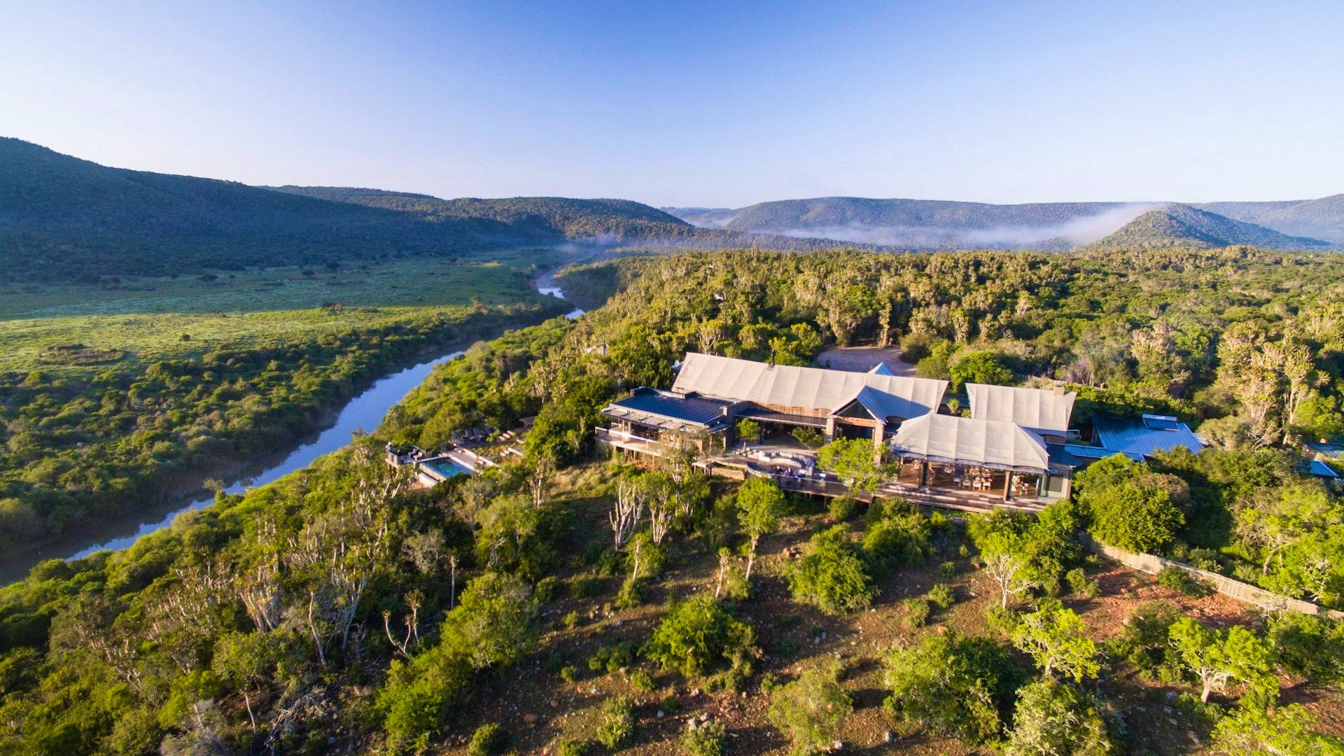 The exterior of a Settlers Drift luxury tent — wooden deck with lounge furniture, canvas roof, freestanding bath, and lush bush all around