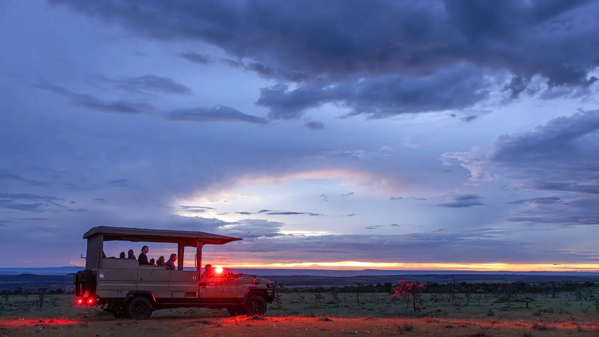 A game drive vehicle full of guests pauses on the open plains at sunset, the sky painted in blues and golds — the end of a perfect safari day